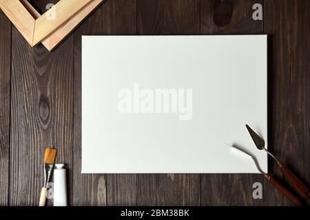 Ein weißer blank grundierter Canvas auf Bahre, Palettenmesser, synthetischer Pinsel und ein Rohr mit Öl- oder Acrylfarbe auf einem braunen Holztisch liegend. Bis Stockfoto