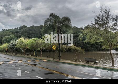 Eine schöne Szene an einem regnerischen Tag, wo man eine leere Bank mit Blick auf einen See sehen kann, sowie einen Regenwald und ein Buckelschild und einen nassen Asphalt. Stockfoto