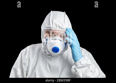 Müde Arzt in weißem Schutzanzug, Gesichtsmaske und Brille. Mann hält den Kopf, Hand in Einweg-blauen Handschuhen, isoliert auf schwarzem Rückgro Stockfoto