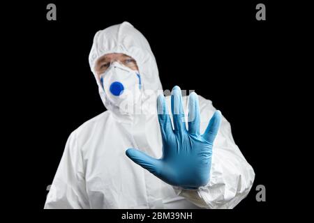 Mann in weißem Schutzanzug mit Stop-Geste, isoliert auf schwarzem Hintergrund. Handschuh blau, Handstoppschild Stockfoto