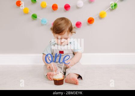 Fröhlicher süßer kaukasischer Babyjunge, der zu Hause seinen ersten Geburtstag feiert. Kind Kleinkind sitzt auf dem Boden im Zimmer. Leckeres Cupcake Dessert mit Cake topp Stockfoto