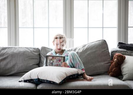 Das blonde Mädchen aus der Schule spielt Tablette auf der Couch mit Teddy Stockfoto