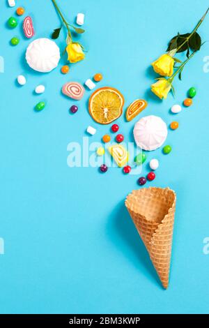 Marshmallows, Süßigkeiten, Gummibärchen, Süßigkeiten, trockenen Becher Orangen und gelben Rosen auf blauem Grund im Wafer Kegel fallen. Textfreiraum Stockfoto