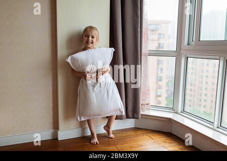 Lächelndes kleines Mädchen in weißen Kissen Kleid zu Hause. Mode Mädchen. Pillow Challenge wegen Aufenthalt. Lustige Quarantäne-Momente während Coronavirus covid-19 Stockfoto