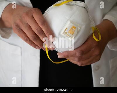 Nahaufnahme einer hispanischen oder Latinx Frauenhand mit einer weißen 3M N-95 Gesichtsmaske mit gelben elastischen Bändern. Stockfoto