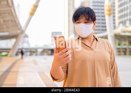 Übergewichtige asiatische Frau mit Maske mit Telefon am Skywalk Brücke Stockfoto