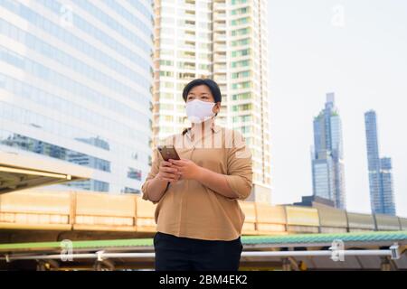 Übergewichtige asiatische Frau mit Maske denken, während Sie Telefon am Skywalk Brücke Stockfoto