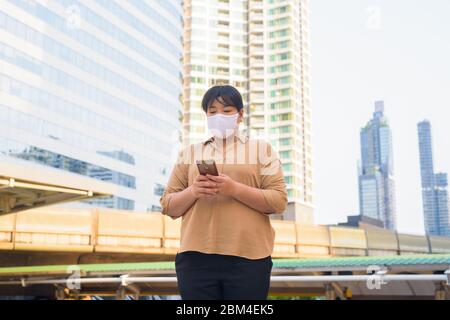 Übergewichtige asiatische Frau mit Maske mit Telefon am Skywalk Brücke Stockfoto