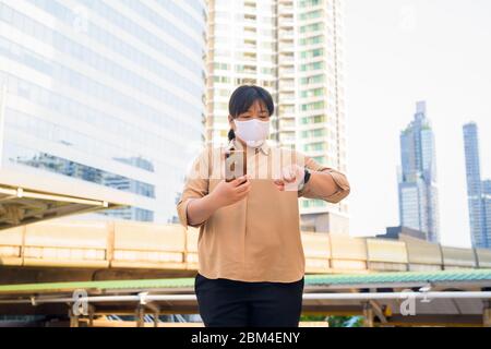 Übergewichtige asiatische Frau mit Maske mit Telefon und Überprüfung der Zeit auf Skywalk Brücke Stockfoto