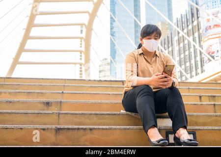 Übergewichtige asiatische Frau mit Maske mit Telefon und sitzen in der Stadt Stockfoto