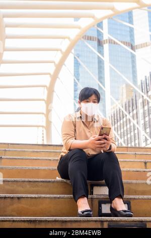 Übergewichtige asiatische Frau mit Maske mit Telefon und sitzen in der Stadt Stockfoto