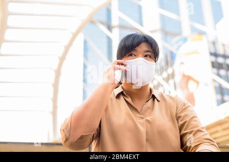 Übergewichtige asiatische Frau mit Maske sprechen am Telefon, während in der Stadt sitzen Stockfoto