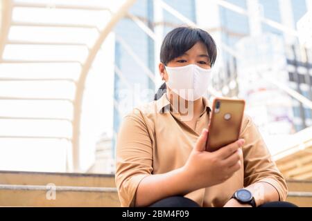 Übergewichtige asiatische Frau mit Maske mit Telefon während in der Stadt sitzen Stockfoto