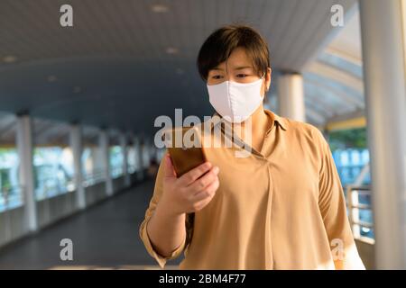 Übergewichtige asiatische Frau mit Maske mit Telefon in der Stadt Stockfoto