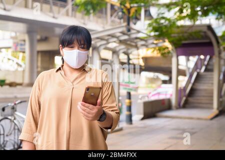 Übergewichtige asiatische Frau mit Maske mit Telefon in der Stadt Straßen im Freien Stockfoto