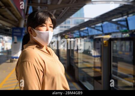 Übergewichtige asiatische Frau mit Maske denken und warten an der Sky-Bahnhof Stockfoto