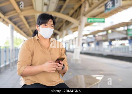 Übergewichtige asiatische Frau mit Maske denken, während Sie Telefon am Sky-Bahnhof Stockfoto