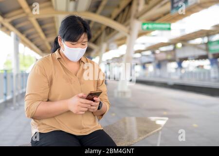 Übergewichtige asiatische Frau mit Maske mit Telefon während sitzen am Sky-Bahnhof Stockfoto