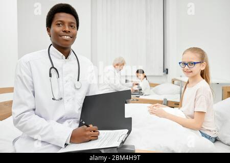 Junger lächelnder afrikanischer Arzt mit Stethoskop am Hals und positivem kleinen Patienten, der im Krankenhaus auf einem Trainer sitzt, Therapeut, der Ordner und Stift hält. Arzt, der den Blutdruck auf dem Hintergrund misst. Stockfoto