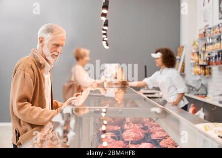 Junge lächelnde Verkäuferin mit lockigen Haaren, die der Dame Würstchen in einer Papierpackung aus dem Kühlschrank gab. Älterer Mann, der auf Glastheke mit großer Auswahl an Fleisch, Supermarkt. Stockfoto