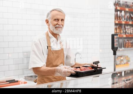 Portrait eines lächelnden männlichen Fleischers, der ein Stück frisches rohes rotes Fleisch in den Händen hält. Fröhlicher Mann hinter der Theke des Ladens, der Fleisch zeigt, die Schüssel aus dem Kühlschrank auf die Waage im Supermarkt setzt. Stockfoto