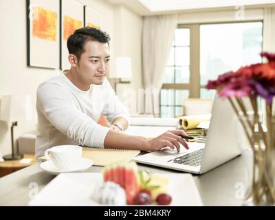 Junge asiatische Geschäftsmann arbeiten von zu Hause sitzen in der Küche Zähler Blick auf Laptop-Computer (Kunstwerk im Hintergrund digital verändert)