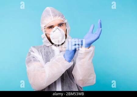 Mann in einem weißen Dekontaminationsanzug, der medizinische Handschuhe auf blauem Hintergrund isoliert anlegt. Coronavirus, Covid-19 und Pandemie-Konzept. Stockfoto