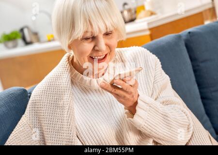 Ruhestand. Ältere Frau zu Hause sitzend, die Sprachnachrichten auf dem Smartphone aufzeichnet und glücklich aus der Nähe lächelt Stockfoto