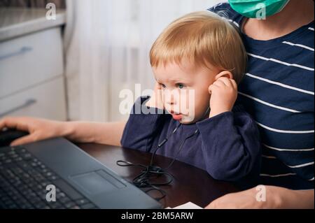 Horizontale Momentaufnahme eines kleinen Helfers, der mit seiner Mutter arbeitet, die während der Quarantänezeit auf dem Schoß am Computer sitzt. Coronavirus Covid-19 Konzept Stockfoto