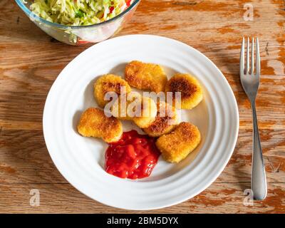 Gekochte Chicken Nuggets aus gefrorenem Produkt (frittiertes paniertes Hühnerfleisch) mit Tomatenketchup auf weißem Teller und Salat aus frischem Gemüse auf altem Stockfoto
