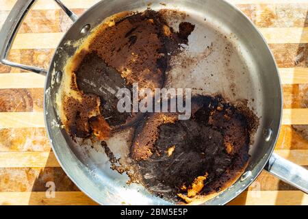 Draufsicht auf Stahlpfanne mit verbrannten Resten beim Kochen auf Holzbrett Stockfoto