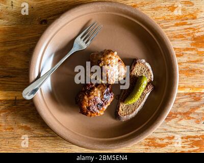 Draufsicht auf zwei gebratene russische Kotleta (panierte Hacksteaks), Roggenbrot und Gurke auf Keramikplatte auf alten Holztisch zu Hause Küche Stockfoto