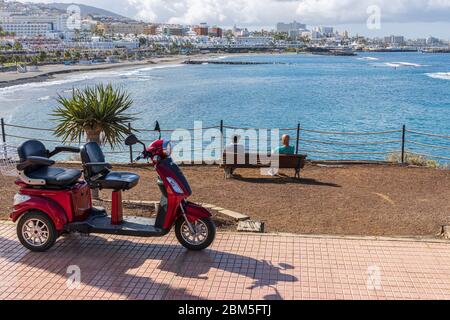 Zwei ältere Männer sitzen auf einer Bank plaudern, während einer ihrer Elektro-Scooter auf der Promenade mit Blick auf Playa Fanabe Strand während der Cov geparkt Stockfoto