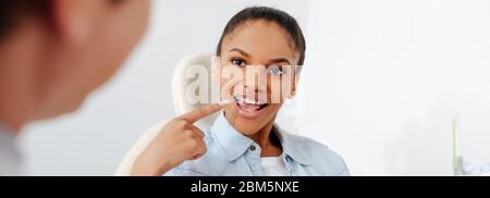 Panoramabild der afroamerikanischen Frau mit geöffnetem Mund, die mit dem Finger auf Zahnspangen in der Nähe des Kieferorthopäden in der Klinik zeigt Stockfoto