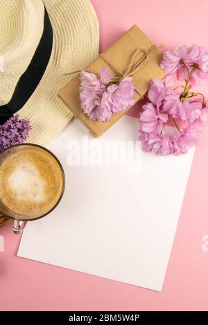 Leeres Papier auf der Zwischenablage, Sommer-Modell. Ein leeres Blatt Papier, umgeben von einem Strandhut, rosa Blumen, Kaffee und Makronen, steht auf einem rosa Stockfoto
