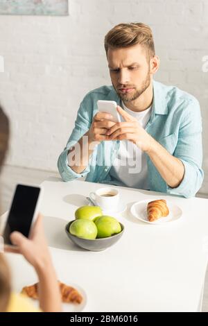 Selektiver Fokus des Mannes mit Smartphone in der Nähe Freundin beim Frühstück in der Küche Stockfoto