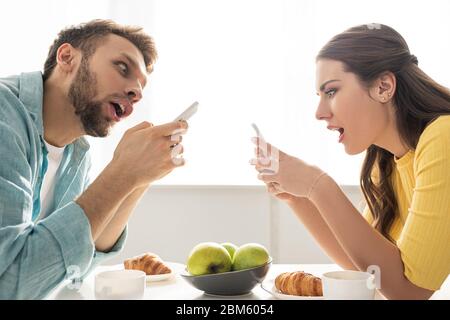 Seitenansicht des schockierten Paar mit Smartphones in der Nähe des Frühstücks auf dem Tisch Stockfoto