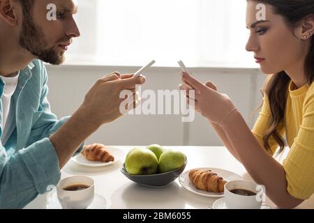 Seitenansicht des Paares mit Smartphones in der Nähe von Kaffee und Croissants auf dem Tisch Stockfoto