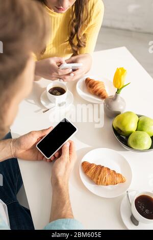 Selektiver Fokus des jungen Paares mit Smartphones in der Nähe von Croissants und Kaffee auf dem Tisch Stockfoto