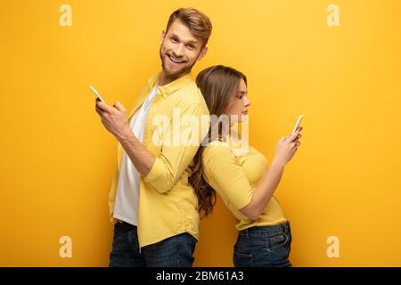 Seitenansicht des Mannes lächelnd auf Kamera in der Nähe Freundin mit Smartphone auf gelbem Hintergrund Stockfoto