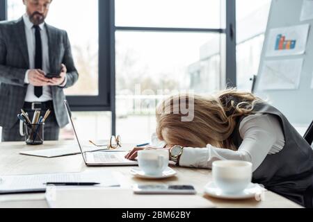 Selektiver Fokus der müden Geschäftsfrau liegt auf dem Tisch in der Nähe von Laptop und Kaffeetassen, während Geschäftsmann mit Smartphone im Büro Stockfoto