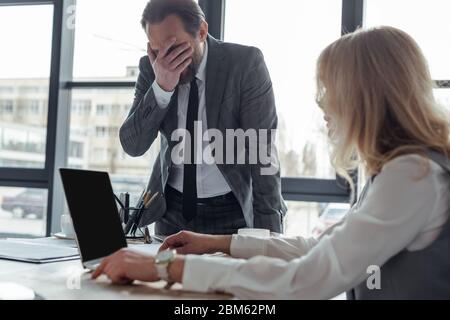 Selektiver Fokus des Geschäftsmannes, der Gesicht mit Hand in der Nähe der Geschäftsfrau mit Laptop bedeckt Stockfoto