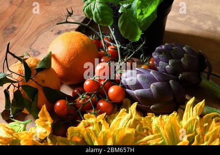 Nahaufnahme auf gelben Kürbisblüten, grünem Basilikum, Orange, Artischocken und roten Tomaten auf Holztisch Stockfoto