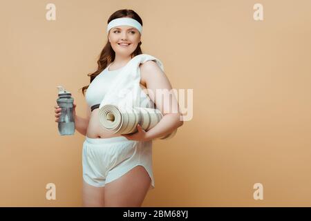 Plus-Größe Frau lächelnd, wegschauen und hält Sportflasche mit Fitness-Matte auf beige Stockfoto