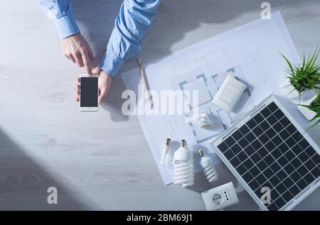 Mann mit einem Mobile touch Bildschirm Smartphone neben einem Solar-Panel, eine Reihe von CFL Lampen und ein Hausprojekt Energiesparkonzept Stockfoto