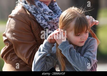 Liebenswert kleines Mädchen binden elastische Bänder auf pinytails auf ihre blonden Haaren im Freien im sonnigen Frühlingspark.Cute Vorschulkinder verbringen Zeit mit ihr m Stockfoto