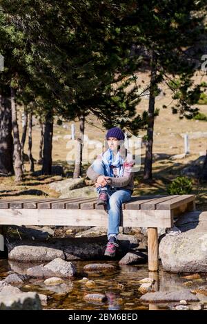 Frau mit einer Wollmütze auf einer Holzbrücke über einen Bach sitzend, während sie in einem Wald sonnenbaden Stockfoto