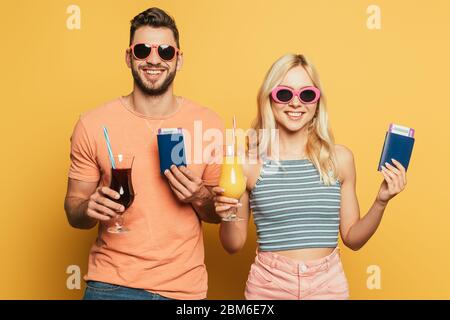 Fröhliches junges Paar in Sonnenbrille hält Cocktails und Dokumente, während lächelnd auf Kamera auf gelbem Hintergrund Stockfoto