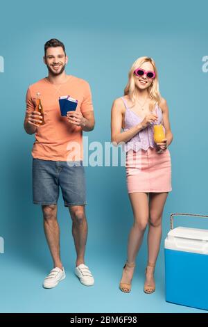 Glücklicher Mann mit Dokumenten und einer Flasche Bier und lächelndes Mädchen, das ein Glas Orangensaft in der Nähe eines tragbaren Kühlschranks auf blauem Hintergrund hält Stockfoto