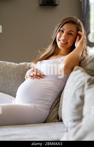 Fröhliche, schöne schwangere Frau, die auf dem Sofa sitzt und ihren Bauch berührt Stockfoto